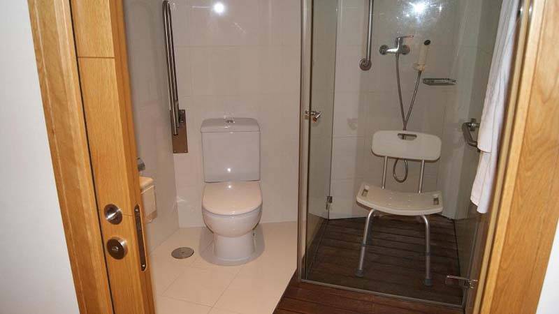Cuarto de baño de la habitación adaptada: inodoro y ducha con barras de apoyo y silla homologada